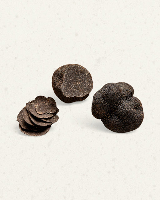 Two whole black winter truffles with a few thin slices fanned beside them, on a light cream background