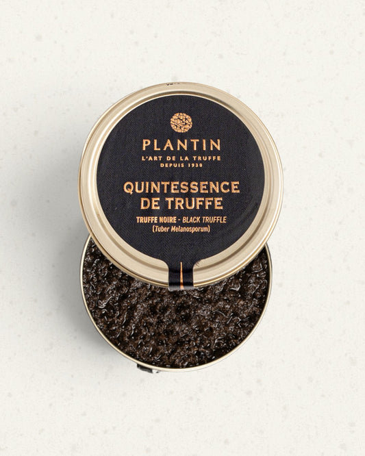 Open black and gold tin of Plantin black truffle quintessence, the lid on the rim above dark truffle pieces