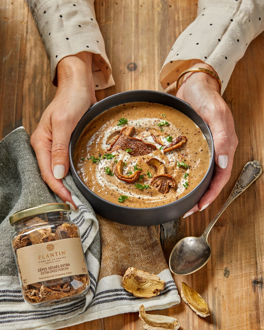 Hands holding a bowl of porcini soup topped with sliced mushrooms, with a jar of dried porcini below