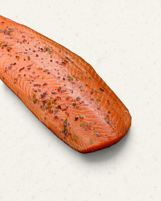 Close-up of a smoked salmon side covered with herbs and pink peppercorns, angled across the frame